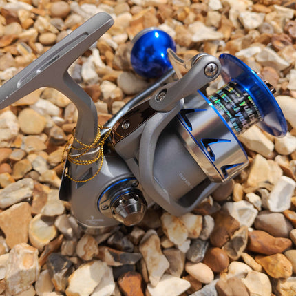 A spinning fishing reel with a metallic gray body, blue accents, and a blue handle knob, photographed resting on small tan and gray pebbles outdoors.