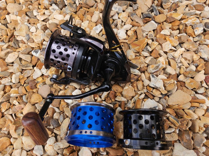 A spinning fishing reel with a black handle and spool, shown alongside two interchangeable spool colors (one blue, one black) on a bed of small tan and brown pebbles.
