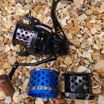 A spinning fishing reel with a black handle and spool, shown alongside two interchangeable spool colors (one blue, one black) on a bed of small tan and brown pebbles.