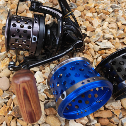 A close-up of a fishing reel with a black body and handle, shown alongside two metal spincast reel bodies in blue and black, placed on a bed of small tan and gray pebbles.