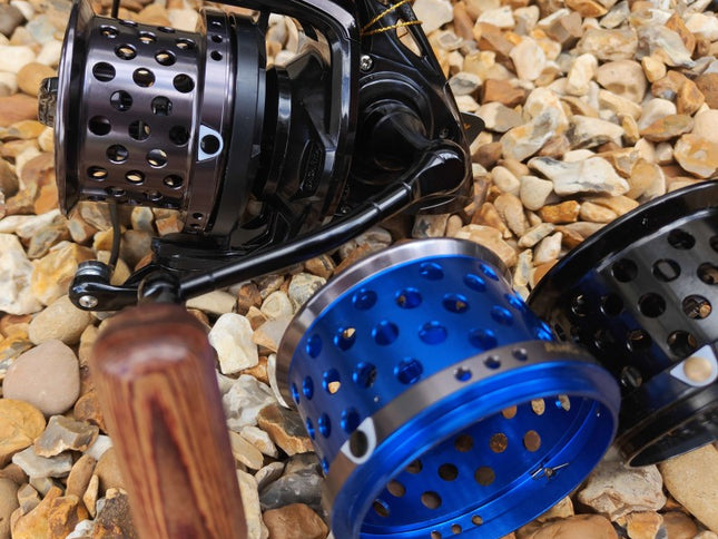 A close-up of a fishing reel with a black body and handle, shown alongside two metal spincast reel bodies in blue and black, placed on a bed of small tan and gray pebbles.