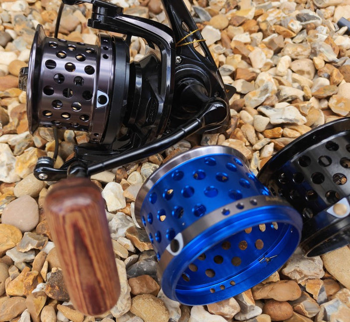 A close-up of a fishing reel with a black body and handle, shown alongside two metal spincast reel bodies in blue and black, placed on a bed of small tan and gray pebbles.