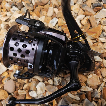 A close-up of a black spinning fishing reel with a perforated metal spool, resting on a bed of small tan and gray pebbles.