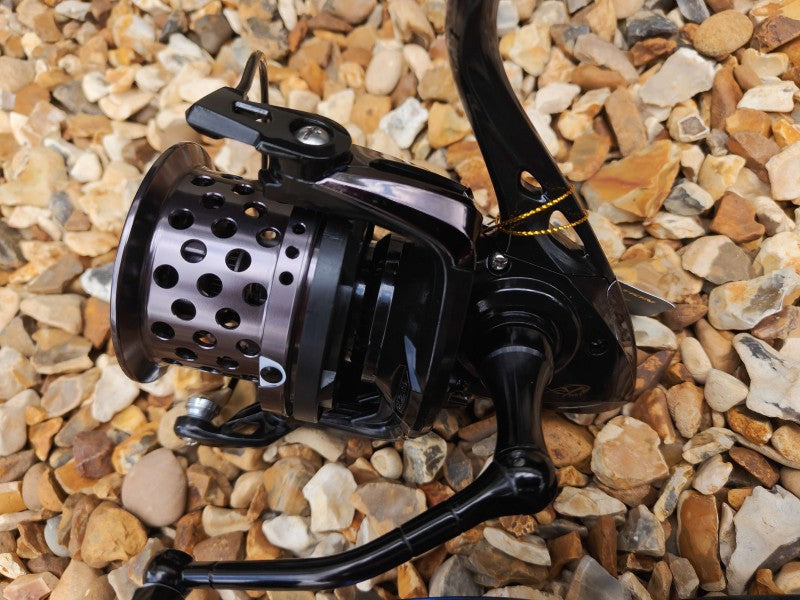 A close-up of a black spinning fishing reel with a perforated metal spool, resting on a bed of small tan and gray pebbles.