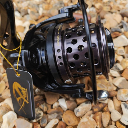 A close-up of a black spinning fishing reel with a perforated metal spool, resting on a bed of small tan and gray pebbles. A black hangtag with a yellow logo is attached to the handle by a yellow cord.