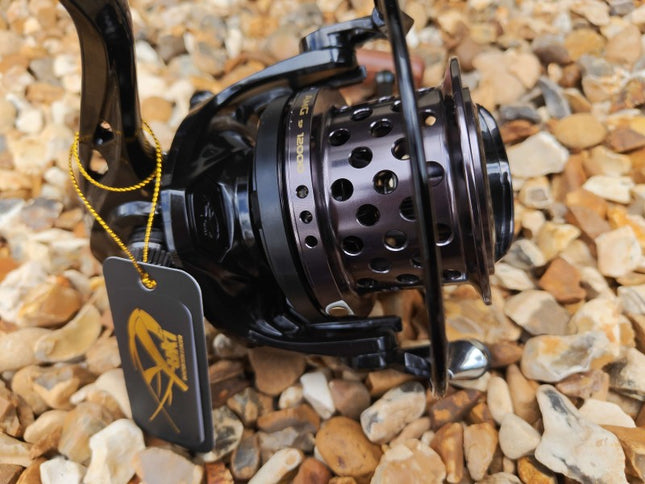 A close-up of a black spinning fishing reel with a perforated metal spool, resting on a bed of small tan and gray pebbles. A black hangtag with a yellow logo is attached to the handle by a yellow cord.