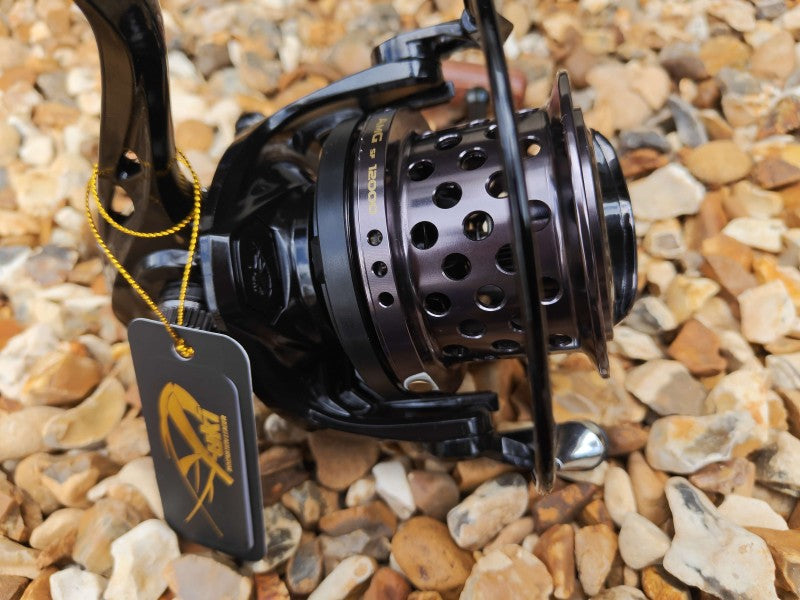 A close-up of a black spinning fishing reel with a perforated metal spool, resting on a bed of small tan and gray pebbles. A black hangtag with a yellow logo is attached to the handle by a yellow cord.