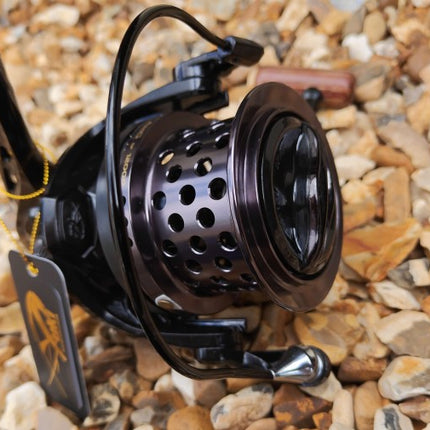 A close-up of a black spinning fishing reel with a perforated metal spool, resting on a bed of small tan and gray pebbles. A small hanging tag is attached to the handle.