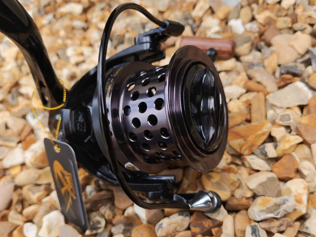 A close-up of a black spinning fishing reel with a perforated metal spool, resting on a bed of small tan and gray pebbles. A small hanging tag is attached to the handle.