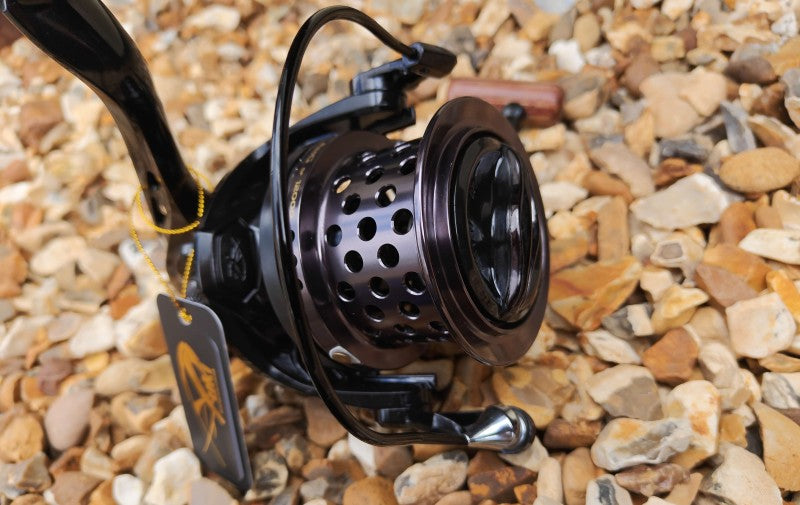 A close-up of a black spinning fishing reel with a perforated metal spool, resting on a bed of small tan and gray pebbles. A small hanging tag is attached to the handle.