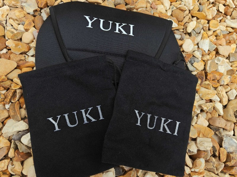 Three black fabric pouches or small bags with the name 'YUKI' embroidered in white on the front, arranged on a bed of small tan and gray pebbles.